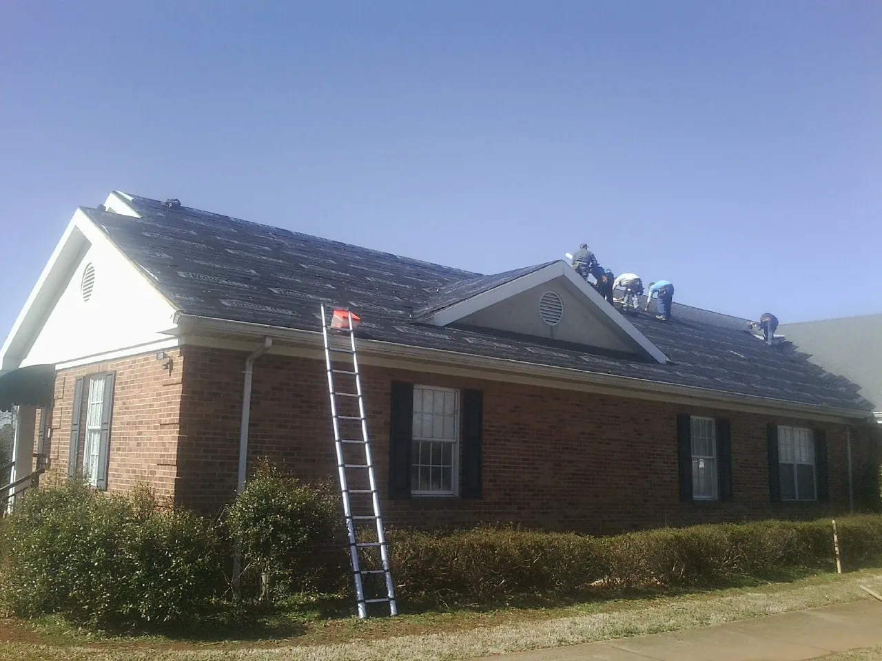 Crew tearing shingles off a brick ranch roof with a ladder set — Metal Roof Repair work by our crew in Excelsior Springs