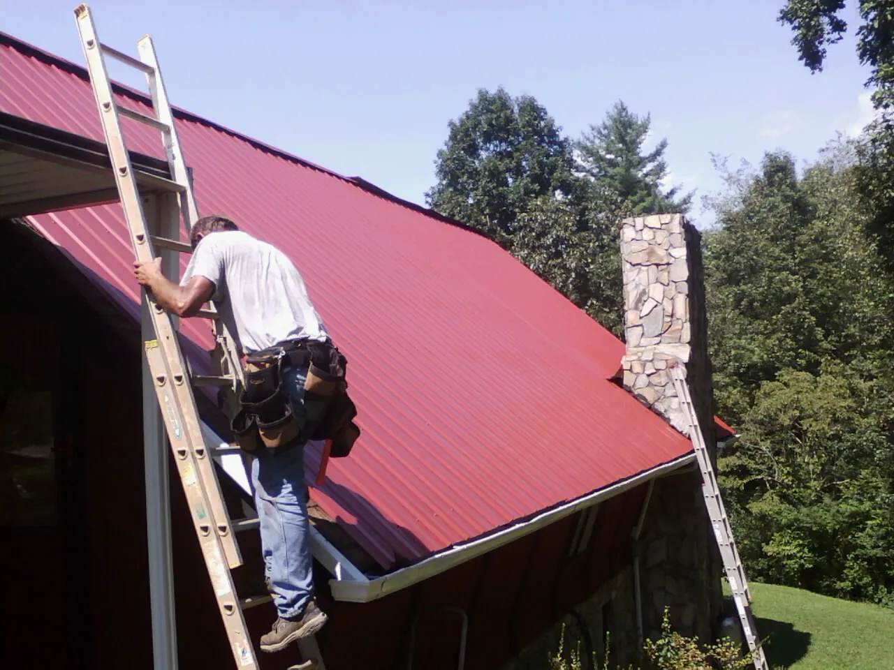 Roofer on a ladder beside a red metal roof and stone chimney — Soffit & Fascia Repair by Roofing Brothers in Excelsior Springs
